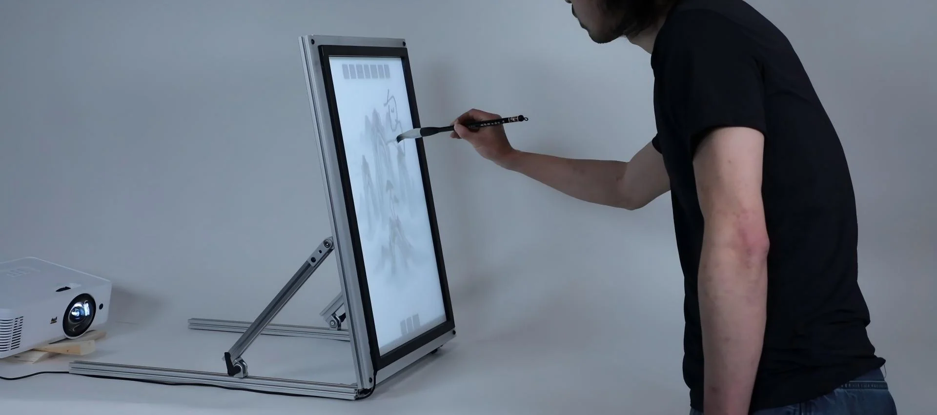 A person writes with a calligraphy brush on a tall back-lit canvas on an aluminium easel; a projector sits on the floor behind it